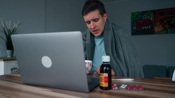 病気の人はノートパソコンでオンラインコースを見てお茶を飲む 男性学生カバー毛布で座っている木製テーブル近くの薬遅い動き — ストック動画