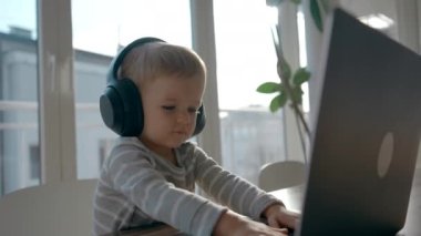 Küçük çocuk kulaklıkla müzik dinleyerek dizüstü bilgisayarın tuşlarına basıyor. Şirin çocuk masada oturmuş modern teknolojiyi yakından inceliyor.