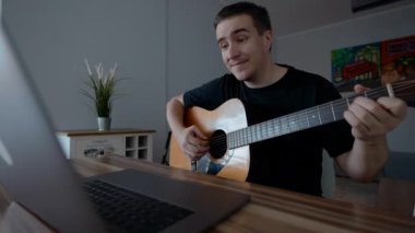 Pozitif adam evde gitar çalmayı öğrenen dizüstü bilgisayarla video dersi izliyor. Genç müzisyen ağır çekimde oturarak yeni beceriler kazanmayı seviyor