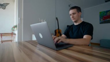 Odaklanmış programcı modern dizüstü bilgisayarda yeni bir video oyunu geliştirir. Alet üzerinde oturan bir adam. Oturma odasındaki tahta masada ağır çekimde oturuyor.