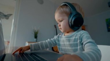 Kulaklıklı tatlı çocuk, babasını taklit eden modern bir dizüstü bilgisayarda yazmaya çalışıyor. Odaklanmış küçük çocuk evdeki elektronik cihazı kullanarak büyük ahşap masada oturuyor.