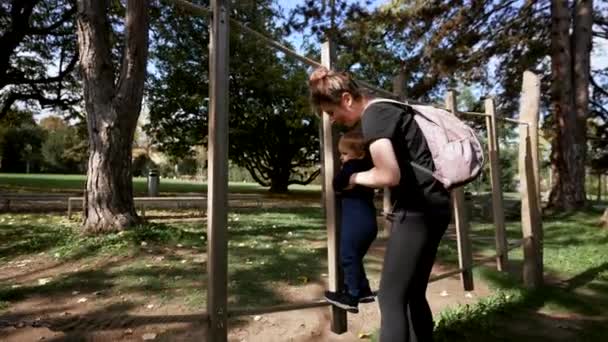 子育ての母親は 日当たりの良い都市公園で木製の構造物の梁に登る間 小さな息子を保持しています バックパックを持つ女性は旅行中にアクティブな幼児を楽しませます — ストック動画