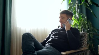 Rahat bir odada rahat bir şekilde dinlenirken, kanepede oturan ve akıllı telefondan konuşan, günlük kıyafetli bir erkek.