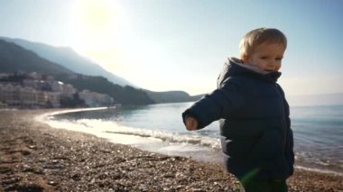 Kumlu kumsalda, dalgalı denizin kıyısında, dağ sırtına ve bulutsuz mavi gökyüzüne bakarken, sıcak giysiler içinde, kameraya bakan küçük şirin çocuğun yan görüntüsü.