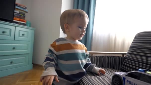 ソファに座り 家で時間を過ごしている間におもちゃを遊んでいるカジュアルな服の小さな男の子の側面図 スローモーションショット — ストック動画