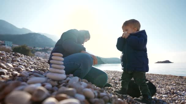母亲和儿子站在宁静的石滩上的卵石塔附近 那男孩把鼻子吹进手帕里 镜头展现了阳光 慢动作的氛围和海滨背景的美丽 — 图库视频影像