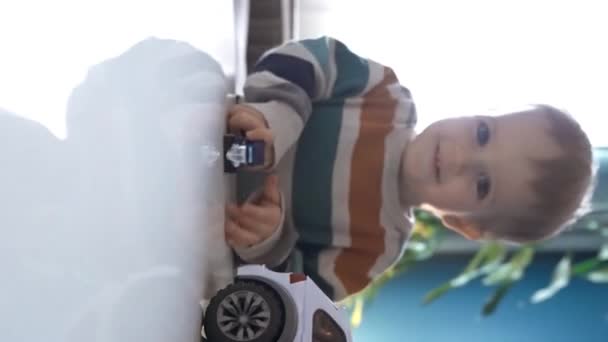 テーブルに座って 家の垂直映像で時間を過ごしながらおもちゃの車を演奏するカジュアルな服の愛らしい少年 — ストック動画
