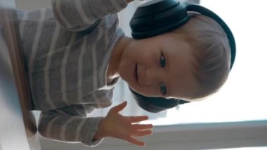 Happy yürümeye başlayan çocuk kulaklıkları ayarlar ve dizüstü bilgisayardan müzik dinler. Meraklı küçük çocuk masada oturmuş modern aletleri izliyor yavaş çekimde dikey görüntüyü kapıyor..