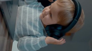 Sevimli küçük çocuk kulaklıkları ayarlıyor kameraya bakıyor ve gülümsüyor. Masum sarışın çocuk, açık dizüstü bilgisayarın yanındaki tahta masada dikey çekimde oturuyor..