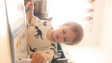 Genç bir çocuk sıcak bir oturma odasında elinde bir diş fırçası tutuyor, kanepede ders veriyor ve kitap okuyor ve bir kalemle egzersiz yapıyor. Dikey görüntüler.