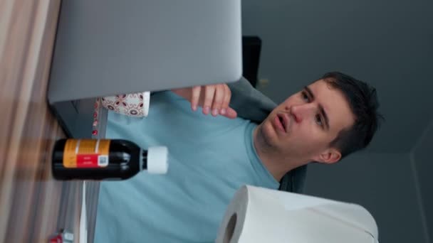 病気のフリーランサーは ノートパソコンで作業中に鼻を拭くためにナプキンをアンロールします 絶望的な男はリビングルームの木のテーブルに座ってインフルエンザを処理しようとします垂直映像 — ストック動画
