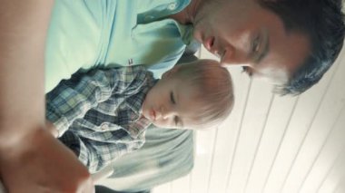 Odaklanmış baba, küçük oğlunu kollarında tutan işaretle çizer. Erkek ve sarışın çocuk, parlak pencereye karşı rahat bir kanepede dinleniyorlar..