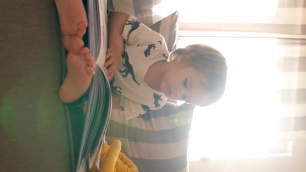 一个小男孩坐在舒适的客厅里的沙发上 一边看书 一边用钢笔做运动 垂直镜头 — 图库视频影像