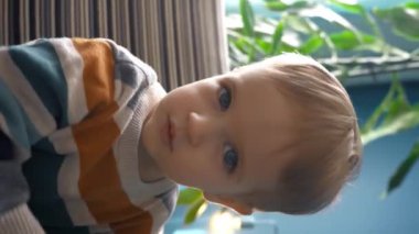 Happy Toddler oturma odasında rahat bir koltukta otururken doğrudan kameraya bakıyor. Geniş açılı yavaş çekim, bu hoş anın sıcaklığını ve çekiciliğini yakalar. Dikey