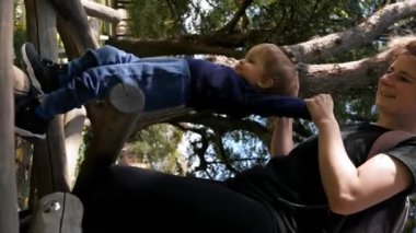 Anne küçük oğlunun elini tutarak tahta kütüklerin arasında yürüyor. Sırt çantalı bir kadın, güneşli bir günde şehir parkında meraklı bir çocuğu eğlendiriyor..