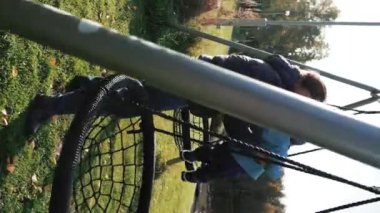 Anne, küçük oğlunu yazlık evin bahçesindeki salıncak yuvasına koymaya çalışıyor. Kadın, güneşli bir sonbahar gününde kırsalda yürüyen bebekle eğlenmekten hoşlanıyor. Yavaş çekim, dikey çekim..