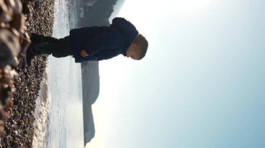 Yürüyen çocuk taşlı sahilde duruyor, denize çakıl taşını yavaş çekimde atıyor. Büyüleyici sahne, dalgalı denizin ve görkemli dağların açık mavi gökyüzüne karşı arka planıyla güçlenir.