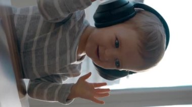 Happy yürümeye başlayan çocuk kulaklıkları ayarlar ve dizüstü bilgisayardan müzik dinler. Meraklı küçük çocuk masada oturmuş modern aletleri izliyor yavaş çekimde dikey görüntüyü kapıyor..