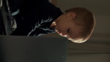 Odaklanmış küçük çocuk, aydınlık pencere ve karanlık köşede yatağında oturan modern dizüstü bilgisayarı inceliyor. Meraklı çocuk evde modern cihazlar kullanıyor. Yakın çekim dikey görüntüler..