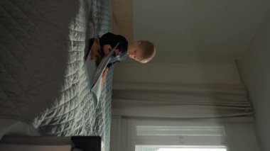 Odaklanmış çocuk yeni kitabı inceliyor sayfaları çeviriyor ve evdeki parlak resimlere bakıyor. Meraklı küçük çocuk yazlık evde yatağında dikey görüntülerde oturuyor..