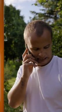 Olgun adam dışarıda telefonda konuşurken rahatlıyor. Beyaz tişörtlü orta yaşlı bir adam özel spor sahasında güneş ışığında dikey çekim yaptıktan sonra bitkin görünüyor..