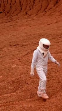 Astronot kraterin içinden geçmeye başlıyor. Beyaz koruyucu giysili ve kasklı bir adam kırmızı gezegenin yüzeyinde yürür, kamera yukarı uçar. Dikey Çekim.