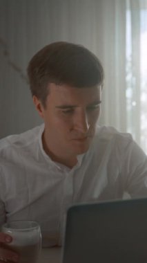 Genç adam evdeki parlak pencereye karşı dizüstü bilgisayara bakarak bardakta kahve içiyor. Adam piyangoda çok para kazanınca şaşırıyor. Dikey çekim..