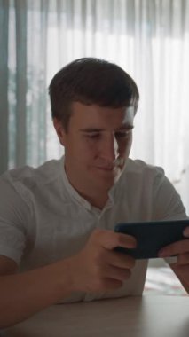 Genç adam akıllı telefonda araba yarışı oynuyor mutfakta parlak pencereye karşı masada oturuyor. Adam evde yakın görünüm dikey çekimde video oyunu seviyor..