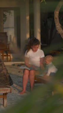 Oğlu ve annesi bir güneş şezlonguna oturup kitap okuyorlar. Hint Okyanusu kıyısındaki kumsal, ağacın arkasından manzara, dikey kamera..