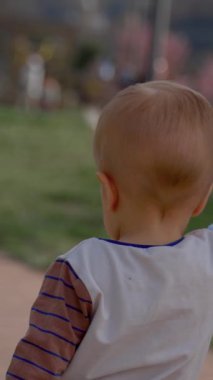 Baba, güneşli bir günde şehir parkında ahşap yolda yürüyen oğlunun elini tutuyor. Bulanık arkaplanda erkek ve çocuk yavaş çekimde dikey çekim.