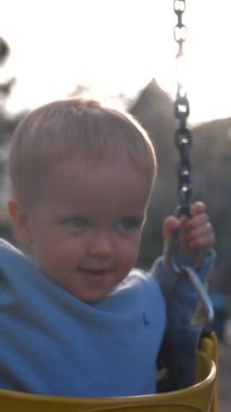 Yürümeye yeni başlayan çocuklar arka planda demir zincirlere tutunarak salıncaklara biniyorlar. Sarışın çocuk, şehir parkında güneşli bir günün keyfini çıkarıyor. Yavaş çekim, dikey çekim..