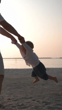 Babasının geniş açılı görüntüsü, çocuğunu okyanus otelinin plajında elleriyle döndürüyor. Yeni yürümeye başlayan bebekle beyaz babasının komik oyunu. Dikey Çekim.