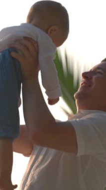 Baba çocuğu kollarına alır ve gökyüzüne fırlatır. Okyanus, büyük bir palmiye ağacı ve arka planda şafak güneşi. Yeni ve rahatlatıcı bir tatil endişesi olmayan bir konsept. Dikey Çekim.