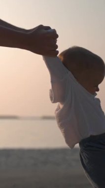 Genç bir baba çocuğunu kumsalda elleriyle çevirir. Küçük çocuklarla eğlenceli bir tatil kavramı. Dikey Çekim.