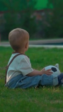 Sarışın çocuk, annesine futbol topu fırlatmak için havuza karşı yumuşak çimenlerde oturuyor. Kadın ve oğul güneşli bir bahar gününün tadını çıkarıyorlar. Ağır çekim dikey çekim..