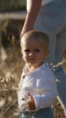 Küçük çocuk sarı çayır çimlerinde duruyor. Annesinin elini tutuyor ve yabani bir çiçek. Sarışın çocuk güneşli bir günde vahşi doğayı keşfediyor. Yavaş çekim dikey çekim..