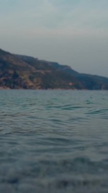 Alacakaranlıkta rüzgarın kumsalları yıkamasından kaynaklanan ışık dalgalarıyla sığ deniz suyu. Bulutlu bir yaz gününde mavi denizin yanındaki orman dağları Dikey Vuruş.