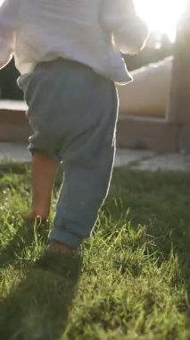 Yürümeye yeni başlayan çocuk, gün batımında evin yakınında büyüyen yeşil çimlerde çıplak ayakla yürümeyi seviyor. Aktif çocuk yaz aylarında avluda ağır çekimde dikey çekim yapar..