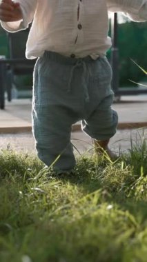 Küçük çocuk güneş ışığıyla aydınlatılmış yeşil çimenlerde yalınayak yürüyor. Şık giyimli küçük çocuk kır evinin bahçesinde vakit geçirmekten hoşlanıyor. Yavaş çekim, dikey çekim..