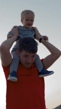 Baba, parlak günbatımında küçük oğlunu sahilde yürürken omuzlarında taşıyor. Adam yaz tatilinde sarışın çocuğu eğlendiriyor. Düşük açı, yavaş çekim, dikey çekim..