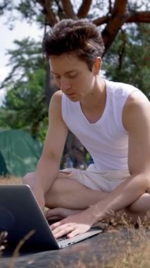 Genç adam, kamp çadırında otururken dizüstü bilgisayarla bilgi yazıyor. Siyah saçlı serbest yazar güneşli bir günde dizüstü bilgisayarda çalışarak temiz havada vakit geçiriyor..