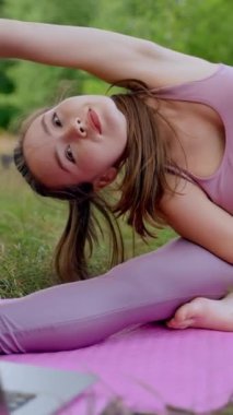 Kahverengi saçlı genç bayan kamp çadırında pilates yapmayı seviyor. Pembe bluz giyen ve tayt giyen bayan turist yoga yapıyor. Kendi dizüstü bilgisayarında dikey çekim yapıyor..