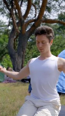 Genç sporcu, çim üzerinde yoga yapıyor. Çimlere karşı çim koyuyor. Çayır üzerinde insanlarla. Siyah saçlı adam yoga yapmayı seviyor. Orman doğasıyla birleşiyor. Yakın plan dikey çekim..