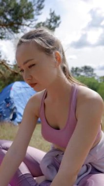 Kahverengi saçlı genç kadın dizüstü bilgisayarda gülümseyen önemli bilgileri yazar. Bayan, dijital cihazlar üzerinde çalışıyor. Temiz havada doğayla bütünleşiyor. Çadır üstüne kurulmuş, dikey çekimde oturuyor..