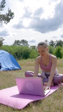 Genç bir kadın dizüstü bilgisayarda çalışıyor, temiz havada kamp çadırında pembe spor minderinde oturuyor. Kahverengi saçlı bayan serbest çalışan, laptopta çalışmaktan zevk alıyor..