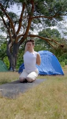 Genç adam çadırın yanında ağaçlara karşı yoga yapıyor. Siyah saçlı yürüyüşçü doğayla bütünleşmek için ellerini uzatıyor. Adam çayırdaki kamp alanında dikey çekim yapmayı seviyor..