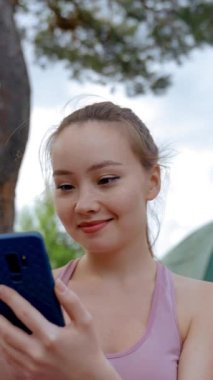 Genç bayan turist internetteki sosyal ağları mavi akıllı telefondan tarıyor. Kahverengi saçlı bayan gülümseyen arkadaşlar gibi görünüyor. Yürüyüşçü doğada dikey çekim yapmaktan hoşlanır..