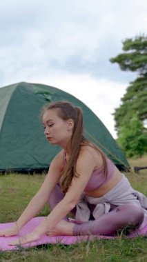 Genç bir kadın pembe paspasın üzerinde bacaklarını esnetir. Kahverengi saçlı kampçı temiz havada yoga yapmaya çalışıyor. Bayan yürüyüşçü, doğada kamp alanında dikey çekim yapmayı sever..