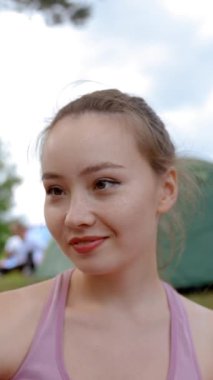 Genç bir kadın telefon kamerasında suratını buruşturarak selfie çekiyor. Kahverengi saçlı bayan kamp alanına karşı çayır doğasında tatil gösteriyor. Kampçı, sağlıklı bir hayat yaşadığını gösteriyor. Yakın çekim dikey çekim..