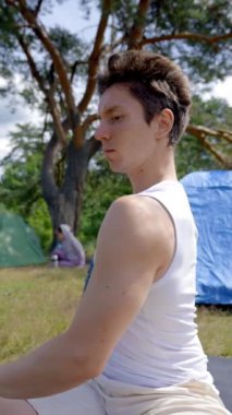 Siyah saçlı yürüyüşçü, orman kamp alanına karşı çimlerin üzerinde yoga yapıyor. Genç adam internetten video izlemenin gerekli kurallarına uyuyor. Dik çekim yakın çekim..